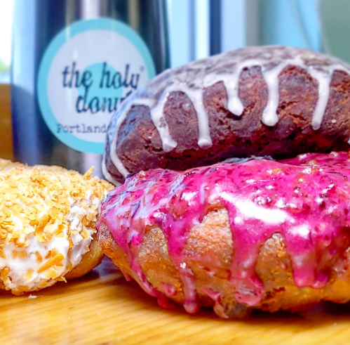 Three colorful donuts: one with pink glaze, one with chocolate icing, and a coconut-covered donut, beside a coffee cup.