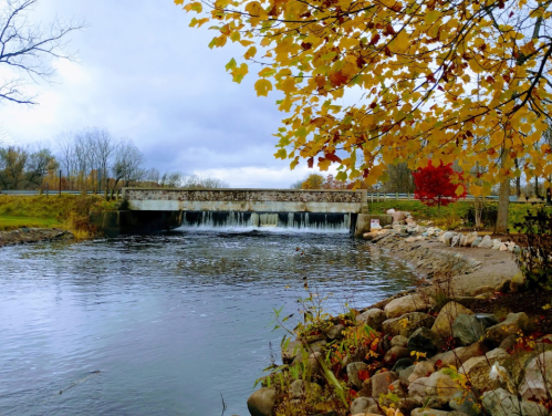 A serene river scene with a small waterfall, surrounded by autumn foliage and a stone-lined bank.