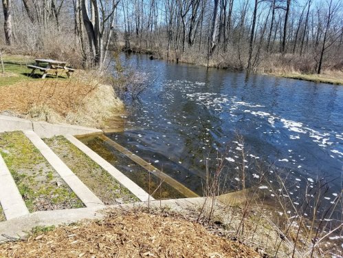 A serene river scene with steps leading down to the water, surrounded by trees and a picnic area in the background.