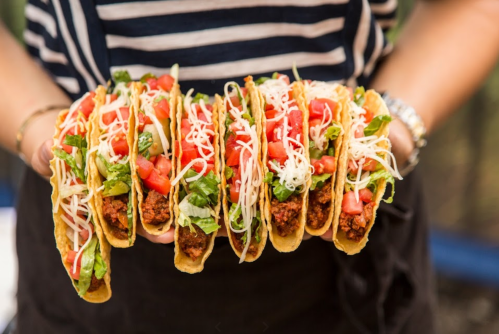 A person holding a platter of six tacos filled with lettuce, tomatoes, cheese, and seasoned meat.