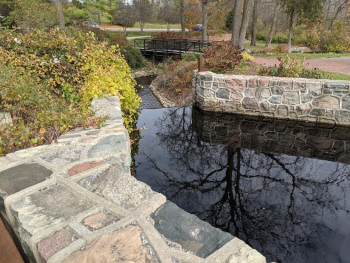 A stone bridge over a dark, reflective waterway surrounded by autumn foliage and trees.