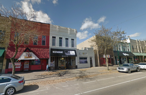 A street view of colorful storefronts, including a restaurant, with trees and a clear blue sky in the background.