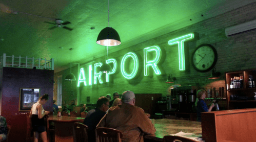 A cozy bar with a neon green "AIRPORT" sign, featuring patrons seated at the counter and a clock on the wall.