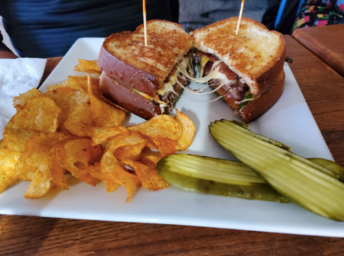 A grilled sandwich with melted cheese and fillings, served with potato chips and pickles on a white plate.