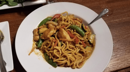 A bowl of stir-fried noodles with chicken, vegetables, and a savory sauce, served with a fork on the side.