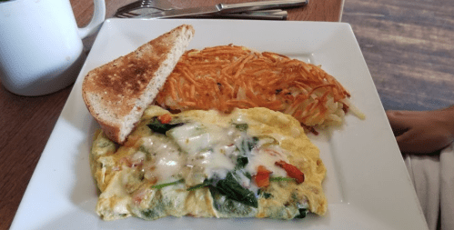 A plate with a vegetable omelette, hash browns, and a slice of toasted bread, served with a coffee mug in the background.