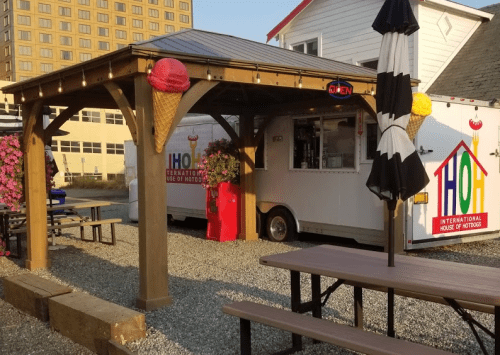 A food truck with a colorful sign under a wooden shelter, surrounded by flowers and picnic tables. Ice cream cones are displayed.