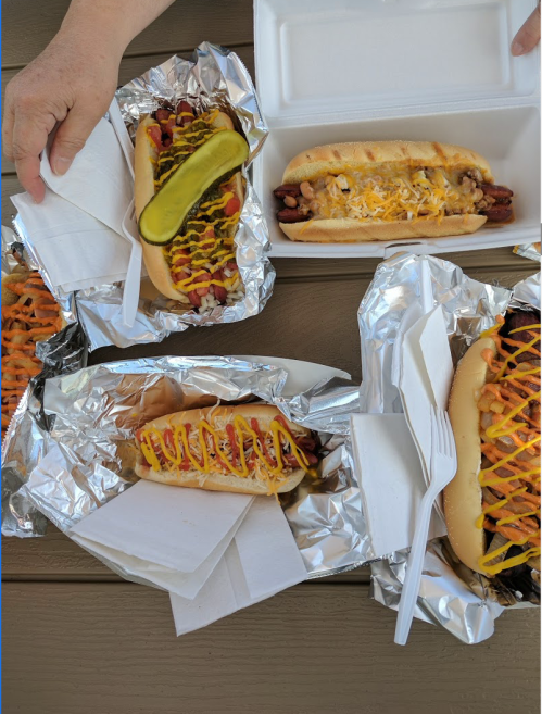 A variety of hot dogs with toppings, served in foil, on a table with a hand holding one.