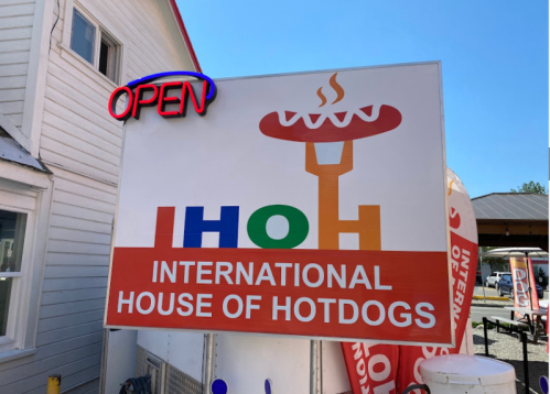 Sign for "International House of Hotdogs" with an open neon sign, featuring a hotdog graphic.