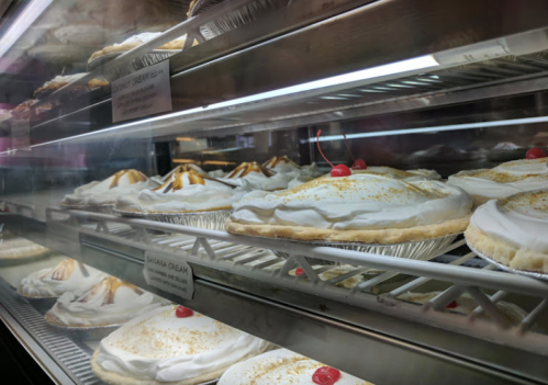 A display case filled with various cream pies topped with whipped cream and cherries.