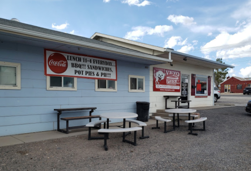 A light blue building with outdoor seating, featuring signs for BBQ, sandwiches, pot pies, and lunch hours.