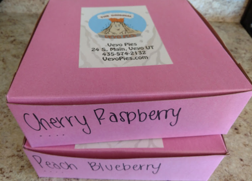 Two pink pie boxes labeled "Cherry Raspberry" and "Peach Blueberry" on a countertop.