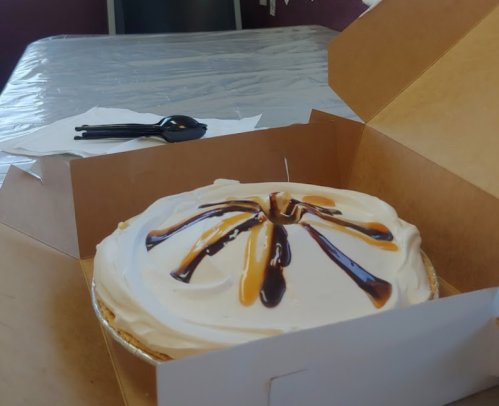 A pie with a creamy topping and drizzled chocolate and caramel sauce, placed in a cardboard box on a table.