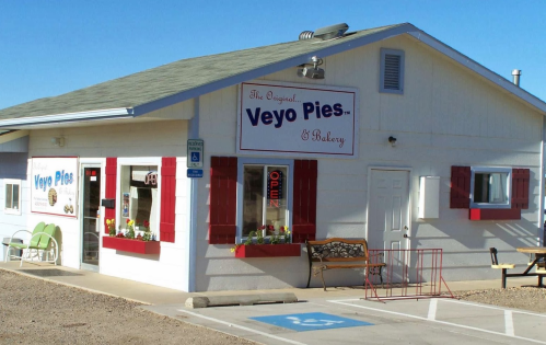 A quaint bakery with a sign reading "Veyo Pies" and red shutters, featuring an open sign and outdoor seating.