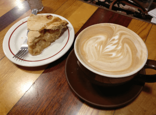 A slice of apple pie on a plate next to a cup of latte with a decorative foam design, on a wooden table.