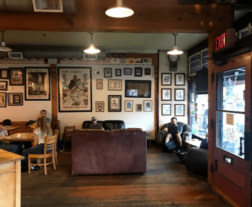 Cozy café interior with wooden floors, framed art on the walls, and patrons sitting at tables and on a couch.