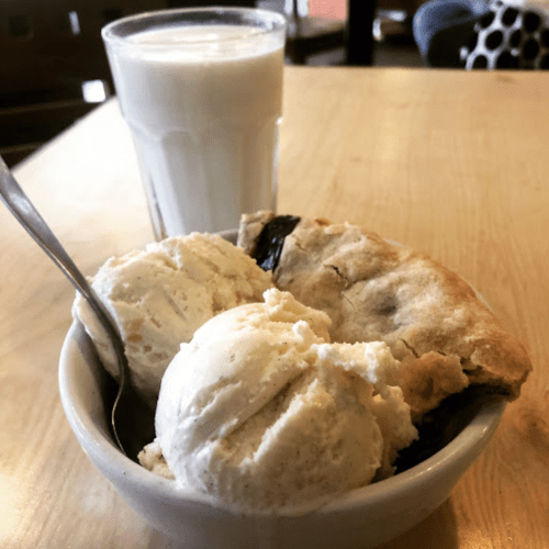 A bowl of vanilla ice cream served with a slice of pie, alongside a glass of milk on a wooden table.