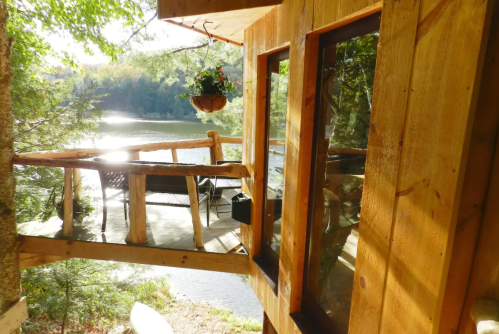 A wooden cabin balcony overlooks a serene lake surrounded by trees, with sunlight reflecting on the water.