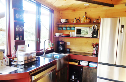 A cozy kitchen with wooden walls, stainless steel appliances, and shelves holding dishes and utensils. Bright natural light.