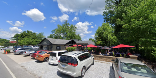 A restaurant with outdoor seating under red umbrellas, surrounded by trees and parked cars on a sunny day.