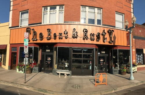 A storefront with the sign "The Bent & Rusty," featuring large windows and a vibrant orange awning.