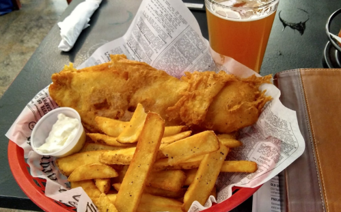 A basket of golden fried fish with crispy fries and a small cup of tartar sauce, alongside a glass of beer.