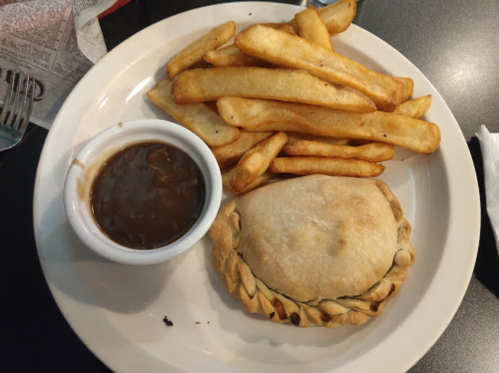 A plate with a baked pastry, fries, and a small bowl of gravy on a black table.