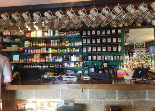 A bar counter with various drinks, snacks, and hanging mugs, featuring shelves stocked with cans and bottles.