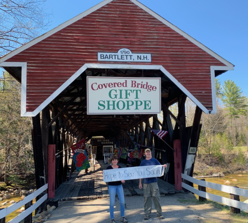 A couple stands outside a covered bridge gift shop, holding a sign that says "Hope to See You Soon!" in Bartlett, NH.