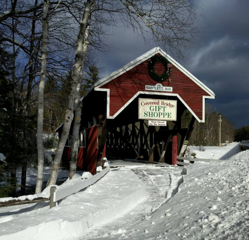 A red covered bridge in Bartlett, NH, surrounded by snow, with a sign for a gift shop. Dark clouds loom overhead.