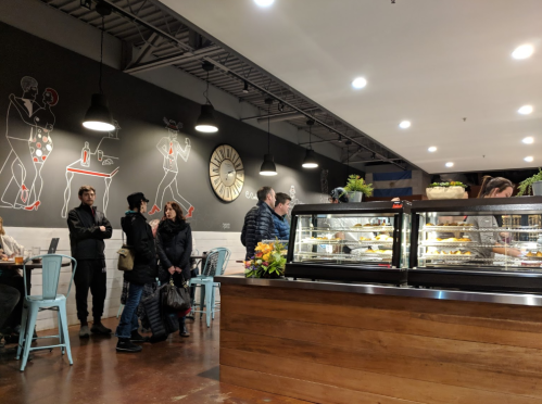 A cozy café interior with a display case of pastries, people chatting, and artistic wall murals.