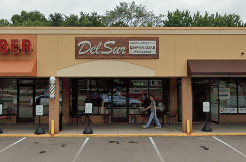 Exterior of Del Sur restaurant featuring a sign for empanadas, with a person walking by and nearby shops visible.