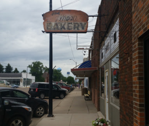 A street view of a bakery sign with a brick building and parked cars under a cloudy sky.