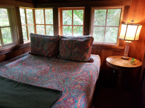 Cozy bedroom with a patterned bedspread, two pillows, a wooden side table, and a lamp, surrounded by large windows.