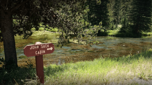 A wooden sign points to "John Sack Cabin" beside a serene pond surrounded by trees and lush greenery.