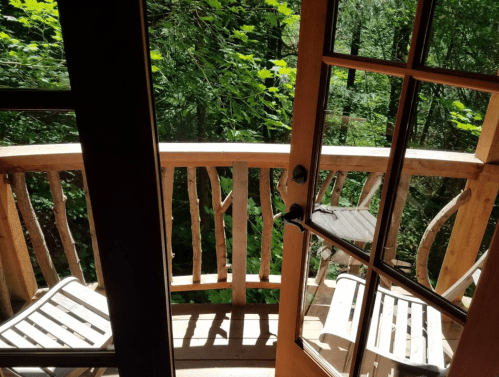 View from an open door to a wooden balcony surrounded by lush green trees. Two chairs are visible on the balcony.
