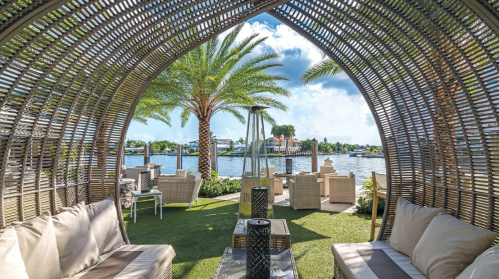 A cozy outdoor lounge with wicker seating, overlooking a serene waterfront and palm trees under a bright blue sky.