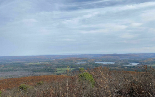 A panoramic view of rolling hills and lakes under a cloudy sky, showcasing autumn foliage in the foreground.