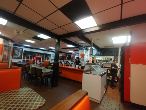 Interior of a diner with orange booths, checkered tables, and staff working in the kitchen area. Brightly lit space.