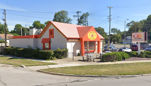 A small diner with a bright orange roof and sign, surrounded by greenery and parked cars on a sunny day.