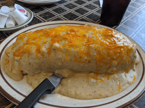 A large, cheesy burrito covered in creamy gravy and topped with shredded cheese, served on a plate.