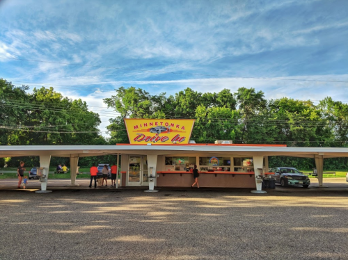 A retro drive-in restaurant with a colorful sign, surrounded by trees and a parking area, on a sunny day.