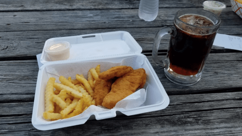 A takeout container with fried chicken strips and crinkle-cut fries, alongside a glass of dark soda on a wooden table.