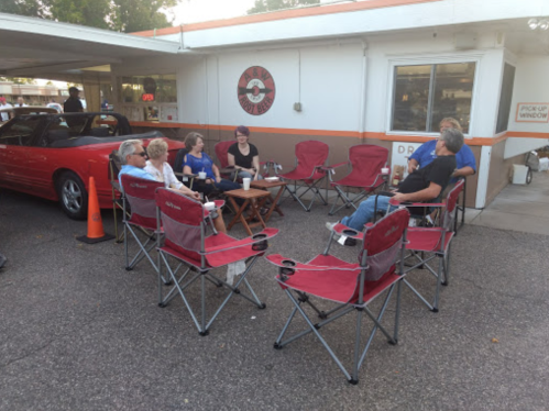 A group of people sitting in red folding chairs outside a building, enjoying drinks and chatting in a casual setting.