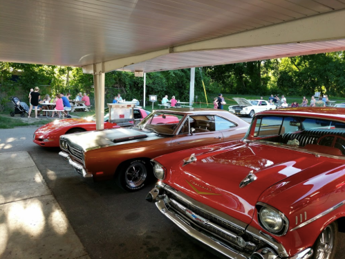 A row of classic cars, including a red convertible and a brown muscle car, parked under a covered area with people in the background.