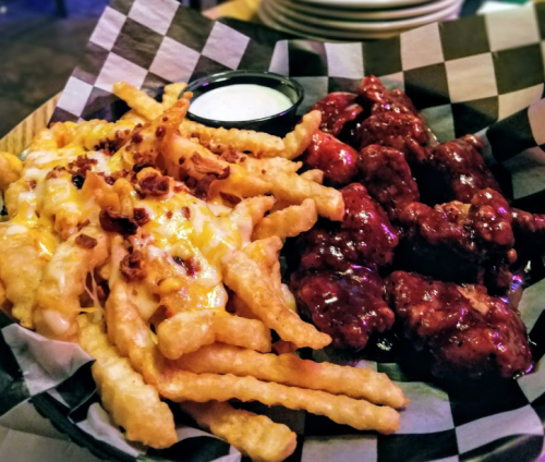 A plate of crispy fries topped with cheese and bacon, alongside saucy chicken wings and a small cup of ranch dressing.