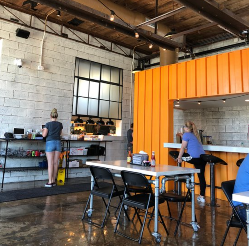 A modern café interior with an orange counter, patrons seated, and a barista preparing drinks in the background.