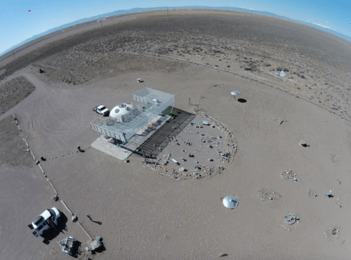 Aerial view of a desert landscape featuring a small building, vehicles, and scattered objects on the ground.