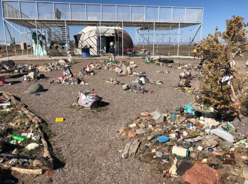 A landscape filled with scattered trash and debris, with a large structure in the background under a clear blue sky.