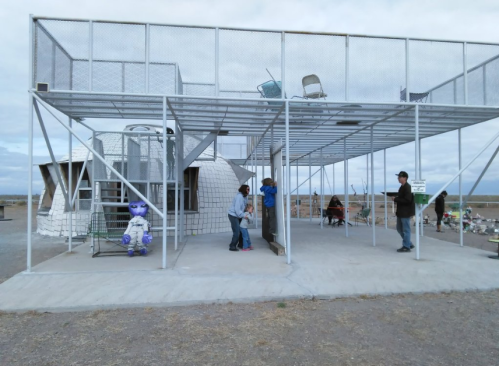 A unique structure with a metal frame, people interacting, and an alien figure nearby on a gravel surface.